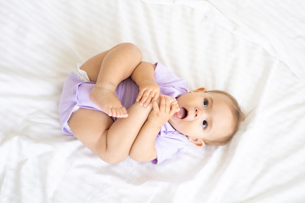 Healthy little cute girl child in bodysuit lies on her back on\
the bed on white bedding plays with her legs the concept of baby\
products