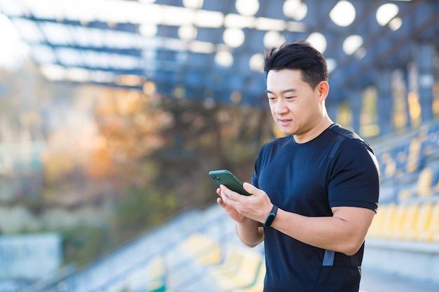 A healthy man watches a video on a mobile phone after a workout while sitting outdoors using an app on a 4g wireless device Asian athlete is happy and smiling after running rest