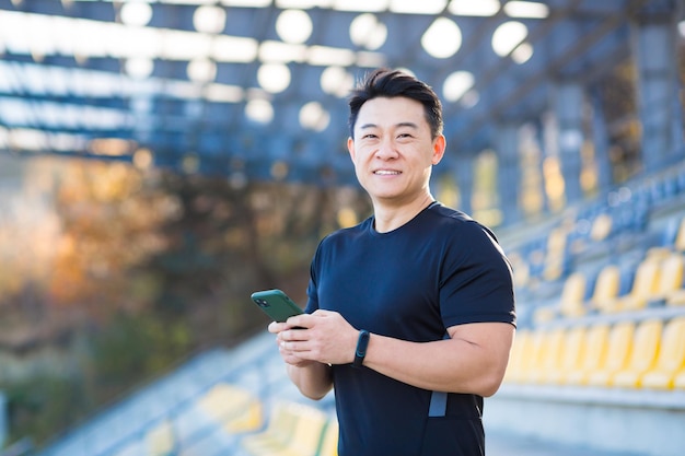 A healthy man watches a video on a mobile phone after a workout while sitting outdoors using an app on a 4g wireless device Asian athlete is happy and smiling after running rest