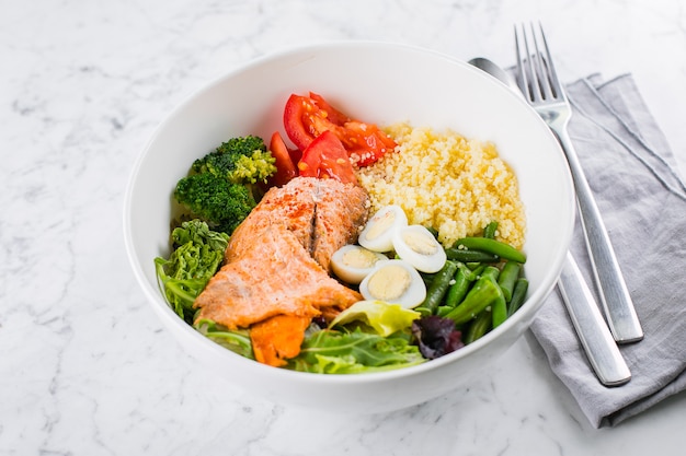 Healthy meal, keto food concept. Fish salad bowl on marble table. Salad with salmon, couscous, vegetables, quail eggs. Top view, copy space