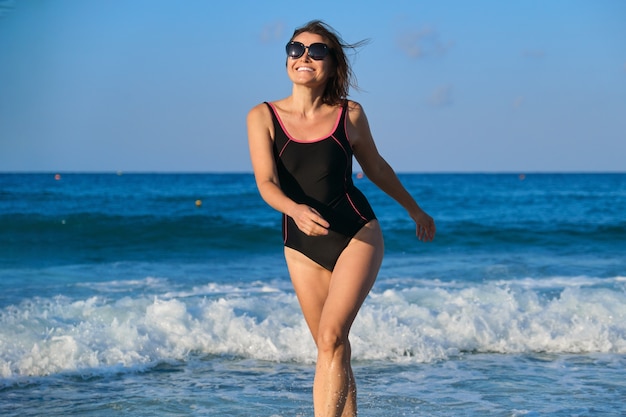 Healthy middle aged woman in sunglasses swimsuit walking along seashore