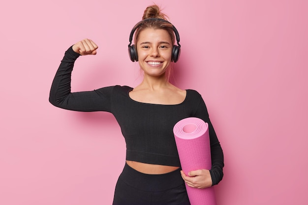 Healthy motivated woman dressed in black sportswear holds rolled karemat listens music via stereo headphones ready for workout isolated over pink background. People sport and health concept.