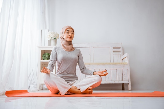 ヒジャブを着た健康なイスラム教徒の女性が、部屋でオレンジ色のマットレスの上でヨガ・ピラティスをしながら瞑想している