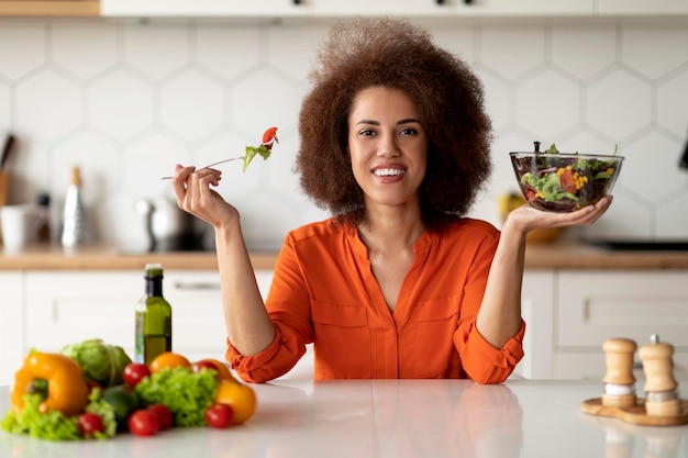 Foto concetto di nutrizione sana felice femmina nera che mangia insalata di verdure fresche in cucina
