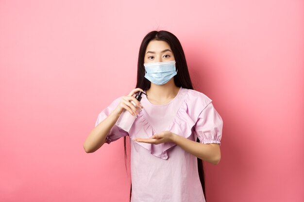healthy people and covid-19 pandemic concept. cute asian woman in medical mask clean hands with antiseptic, using sanitizer, standing against pink background.
