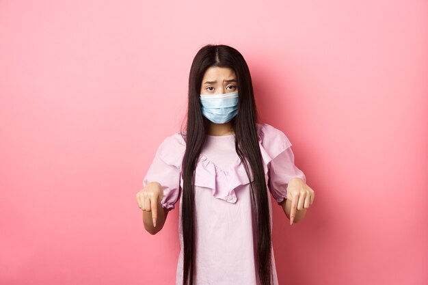 Healthy people and covid-19 pandemic concept. Sad asian woman pointing fingers down, frowning upset, standing against pink background.