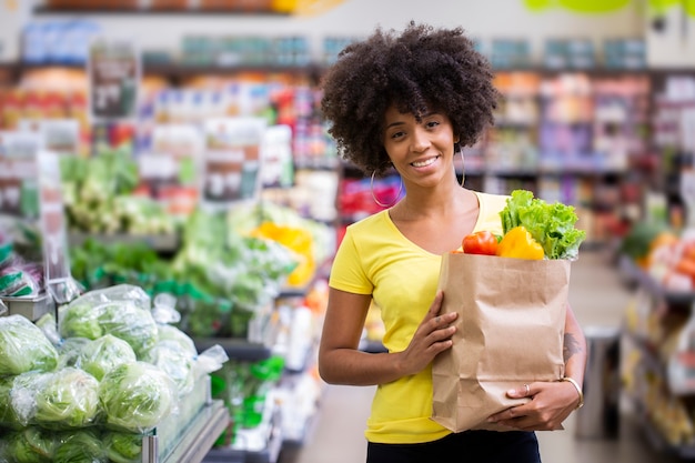 Donna africana felice positiva sana che tiene un sacchetto della spesa di carta pieno di frutta e verdura.