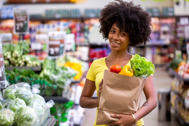 果物や野菜でいっぱいの紙の買い物袋を持っている健康でポジティブな幸せなアフリカの女性。