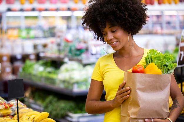 果物や野菜でいっぱいの紙の買い物袋を持っている健康でポジティブな幸せなアフリカの女性。