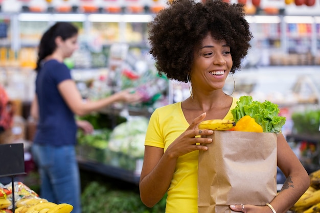 果物や野菜でいっぱいの紙の買い物袋を持っている健康でポジティブな幸せなアフリカの女性。