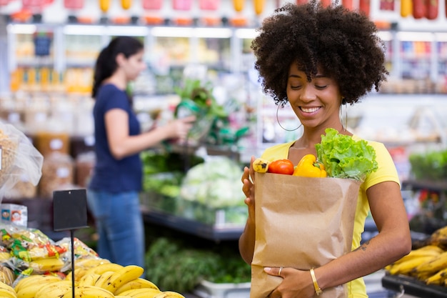 果物や野菜でいっぱいの紙の買い物袋を持っている健康でポジティブな幸せなアフリカの女性。