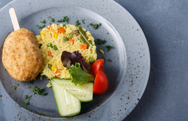 Healthy restaurant food background. Vegetable risotto, chicken cutlet served with fresh cucumber and tomato on grungy blue plate on gray table. Organic everyday meals closeup