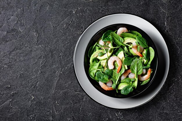 Healthy salad of avocado shrimps with baby spinach leaves, fresh sunflower sprouts, olive oil dressing, and chili flakes served on a plate on a dark background, top view, close-up, copy space