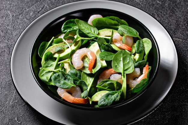 Healthy salad of avocado shrimps with baby spinach leaves, fresh sunflower sprouts, olive oil dressing, and chili flakes served on a plate on a dark background with cutlery, top view, close-up