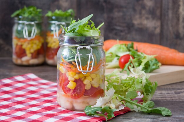 Полезный салат в масонской банке с белой фасолью, помидорами, луком, кукурузой и салатом на деревянном столе