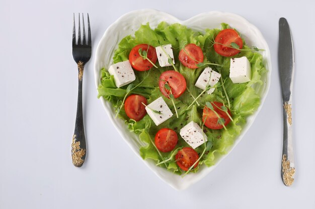 Healthy salad with cherry tomatoes, feta and peas microgreens on a heart-shaped plate on a white table, healthy eating day, Top view