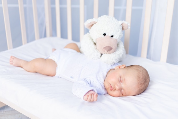 Healthy sleep of a newborn baby in a crib in the bedroom with a soft toy bear on a cotton bed.