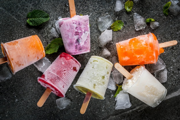 Healthy summer desserts. Ice cream popsicles. Frozen tropical juices, smoothies blueberries. currants, orange, mango, kiwi, banana, coconut, raspberry. On black stone table, plate copyspace top view