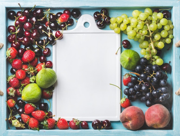 Una sana varietà di frutta estiva fichi uva nera e verde ciliegie fragole pesche su sfondo di legno dipinto di blu con bordo in ceramica bianca nello spazio centrale della copia