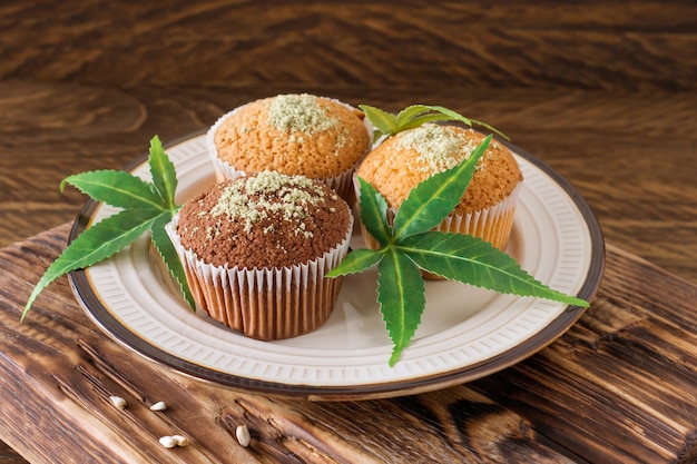 Healthy and tasty vegan and gluten free muffins topped with hemp seeds on a white plate on wooden table. Marijuana cupcake muffins with cannabis leaves.