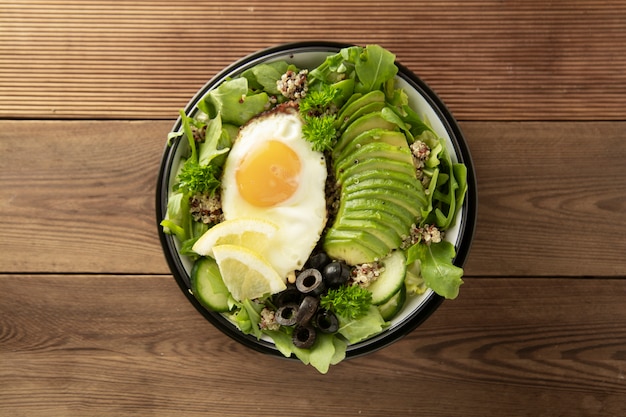 Healthy vegetarian breakfast bowl. Quinoa, avocado, egg, green salad, black olives over wooden background. Energy boosting, clean eating, diet food concept