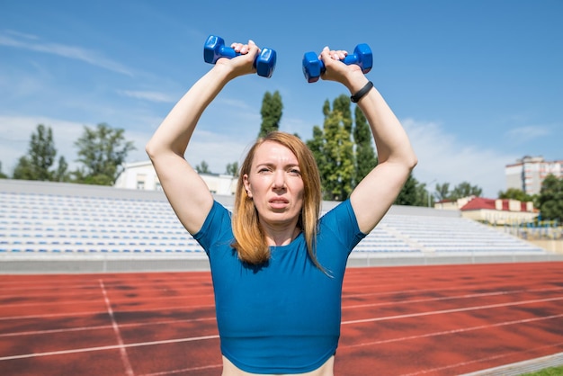 Healthy woman exercising in the stadium with small blue dumbbells Exercising with dumbbells outdoors Runner training before fitness and jogging in the park The concept of a healthy lifestyle