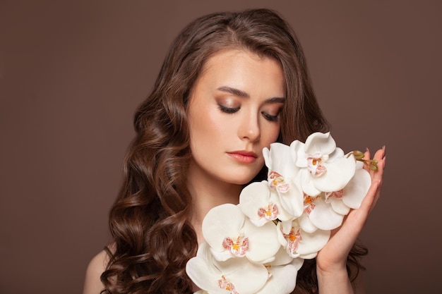 Healthy woman with clear skin brown curly hairstyle and white orchid flowers Facial treatment hair and skin care concept