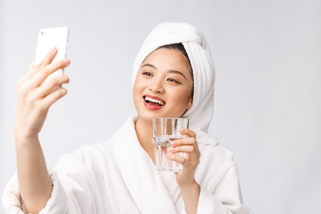 Acqua potabile della giovane bella donna in buona salute, trucco naturale del fronte di bellezza con la tenuta del telefono cellulare, isolato più