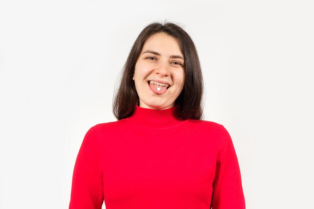 A healthy young woman in red with pills on her tongue smiles and looks into the camera on a white background