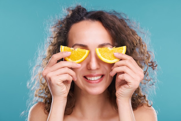 Healthy young woman with toothy smile and dark wavy hair holding two halves of juicy orange slice by her eyes during beauty procedure