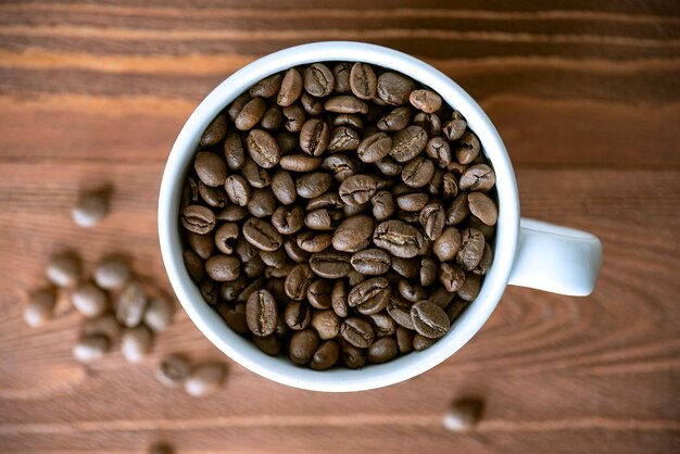 Heap of coffee beans in white ceramic  mug on wooden table. Roasted coffee grains on wooden surface, top view