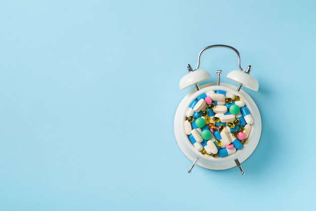 Heap of colorful pills and capsules and alarm clock on blue. Time and healthcare concept. top view