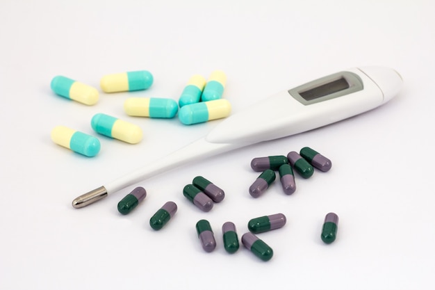 Heap of deifferent pills and digital thermometer on white backgroundg. Dark green and lilac, light yellow and blue cupcules