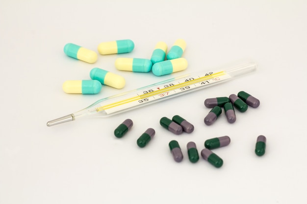 Photo heap of deifferent pills and mercury thermometer on white backgroundg. dark green and lilac, light yellow and blue cupcules