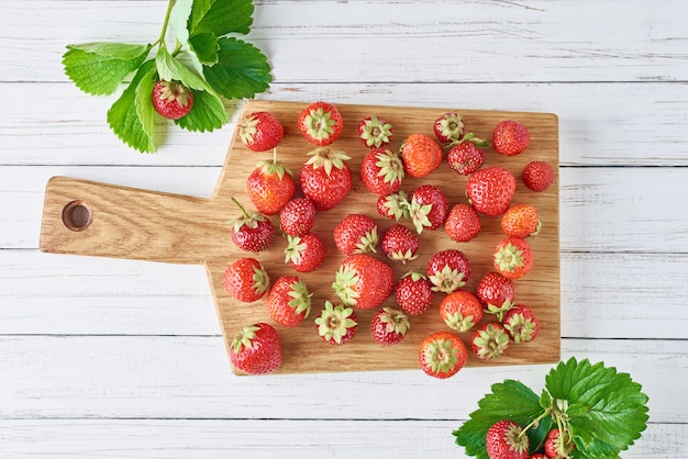 Heap of fresh strawberries with cutting board on a white wooden table, top view. Healthy nutrition