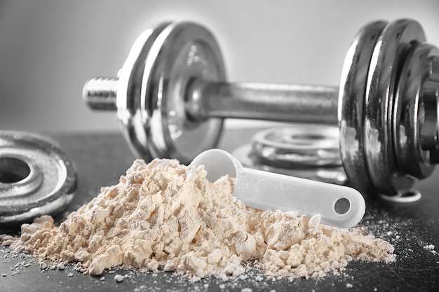 Heap of protein powder with scoop and dumbbell on table closeup