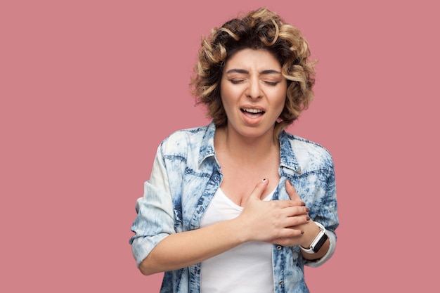 Photo heart attack or pain. portrait of sick young woman with curly hairstyle in casual blue shirt standing and holding her painful herat and feeling bad. indoor studio shot, isolated on pink background.