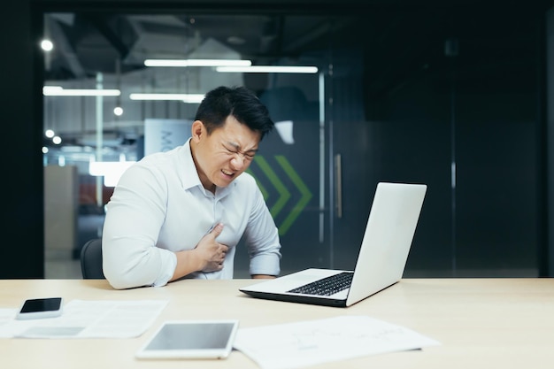 Heart attack at work a young asian man businessman office worker holds his heart feels pain had a