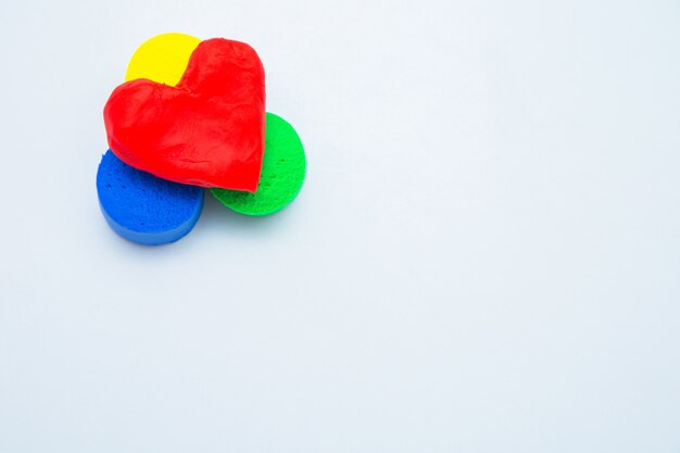 A heart made of red dough for modeling lies on top of a yellow green and blue mug on a white backgro...