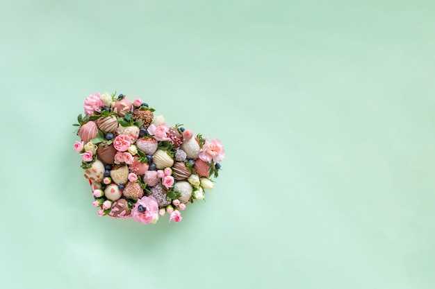 Heart shaped box with handmade chocolate covered strawberries with different toppings and flowers as a present on Valentines day on green background with free space for text