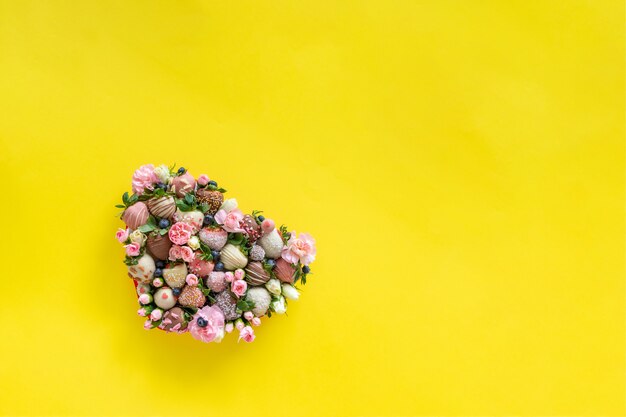 Heart shaped box with handmade chocolate covered strawberries with different toppings and flowers as a present on Valentines day on yellow background with free space for text