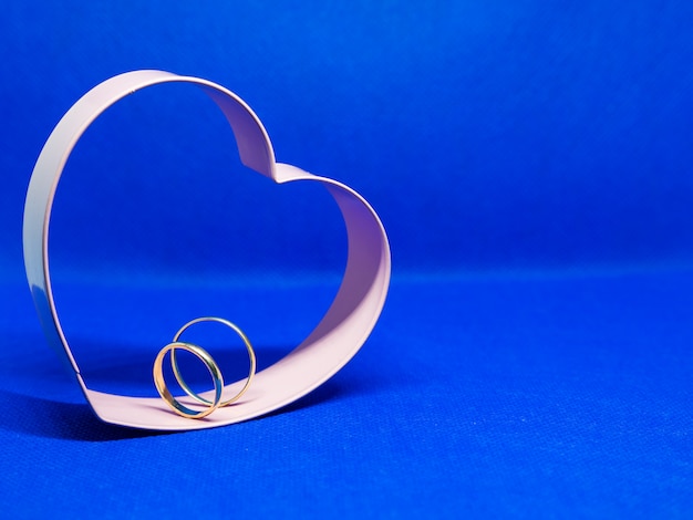 Heart-shaped cookie mold frame. In the center wedding rings. Blue background, isolated, copy space for message. Valentine's day concept Declaration of love.