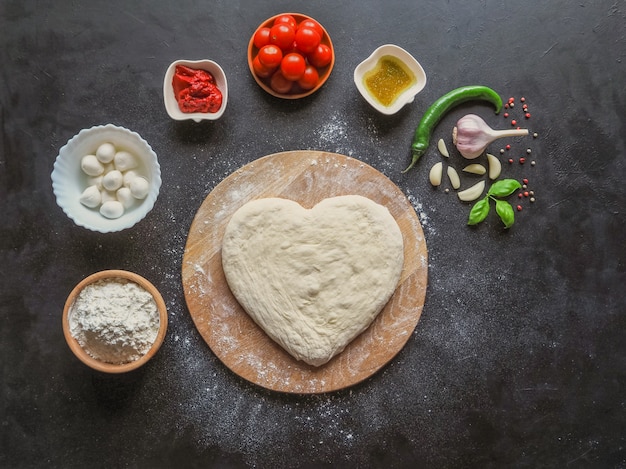 Heart-shaped dough and a set of ingredients for pizza on a black table. The view from the top