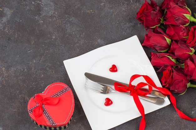подарочная коробка в форме сердца, тарелка со столовыми приборами, красные сердечки и букет красных роз