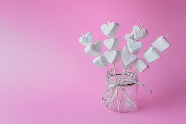 Heart shaped marshmallows on wooden sticks on pink paper background in glass jar