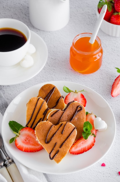 Heart-shaped pancakes with chocolate sauce and fresh strawberries on a white plate with a cup of coffee