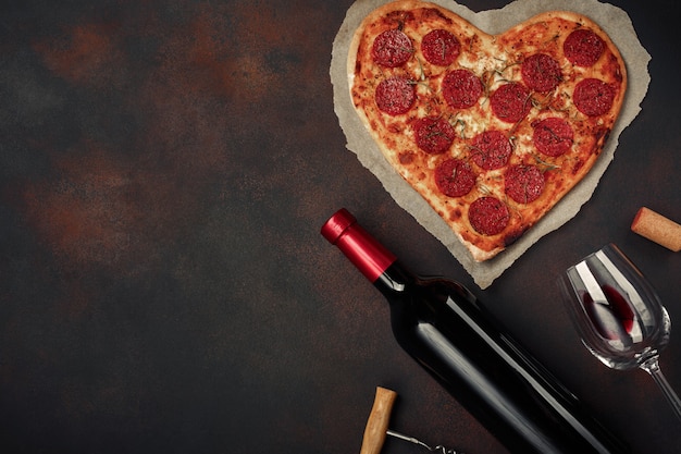 Heart shaped pizza with mozzarella, sausagered with a bottle of wine and wineglas on rusty background.