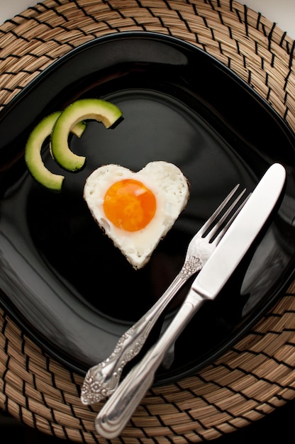 Heart-shaped scrambled eggs with avocado on a black plate on a\
jute napkin with cutlery