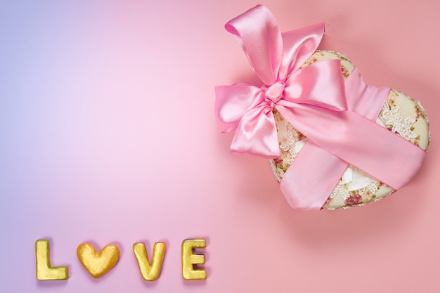 Heart shaped Valentines Day gift box with pink curved ribbon and gold word love on paper background.