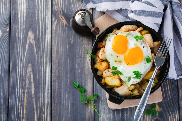 풍성한 아침 식사. 갈색 감자, 닭고기, 양파, 파슬리, 오레가노 및 튀긴 계란을 오래된 나무 테이블에있는 프라이팬에 해시합니다. 평면도